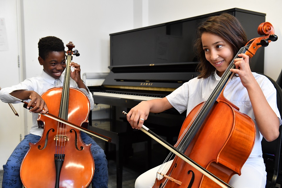 A pair of young students, performing on the cello, smiling at each other, with a piano in the background.